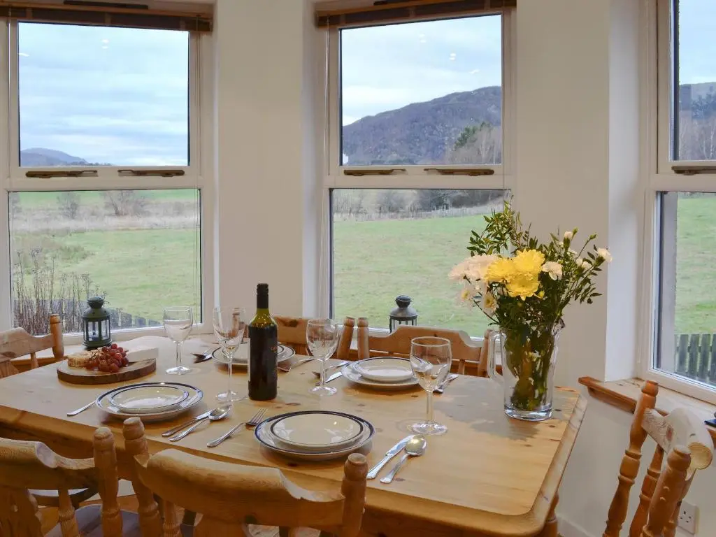 a dining room table with a bottle of wine and windows at Birch Tree Cottage in Aviemore