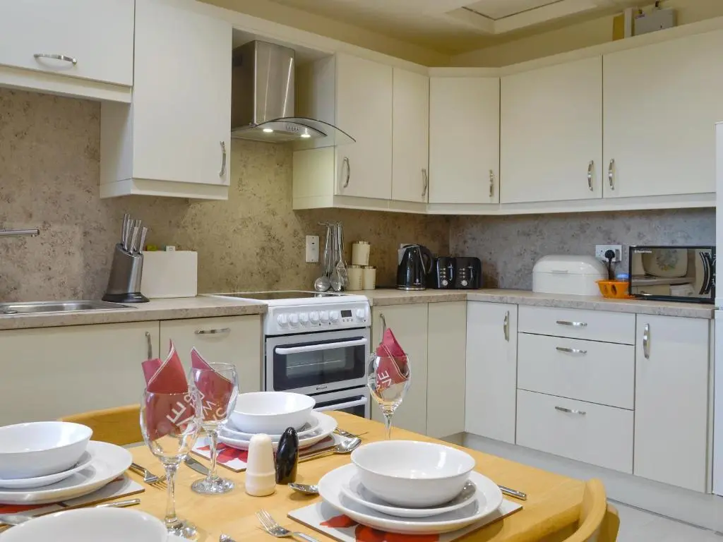 a kitchen with white cabinets and a table with plates and glasses at Mill Shore Cottage - 26817 in Pennan
