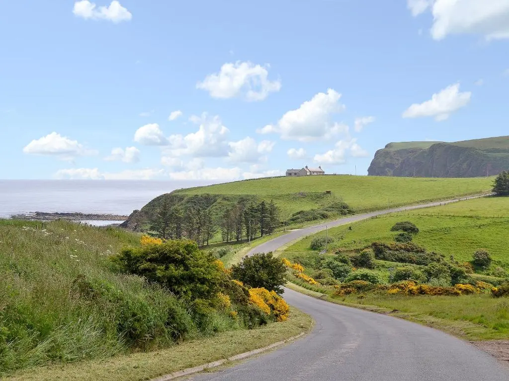 a winding road on a hill with a house in the distance at Mill Shore Cottage - 26817 in Pennan