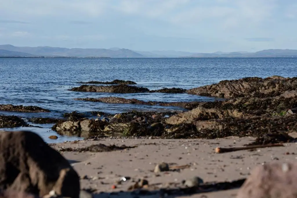 a beach with rocks and water with mountains in the background at Ina's Cottage - Seasgair Lodges in Portmahomack