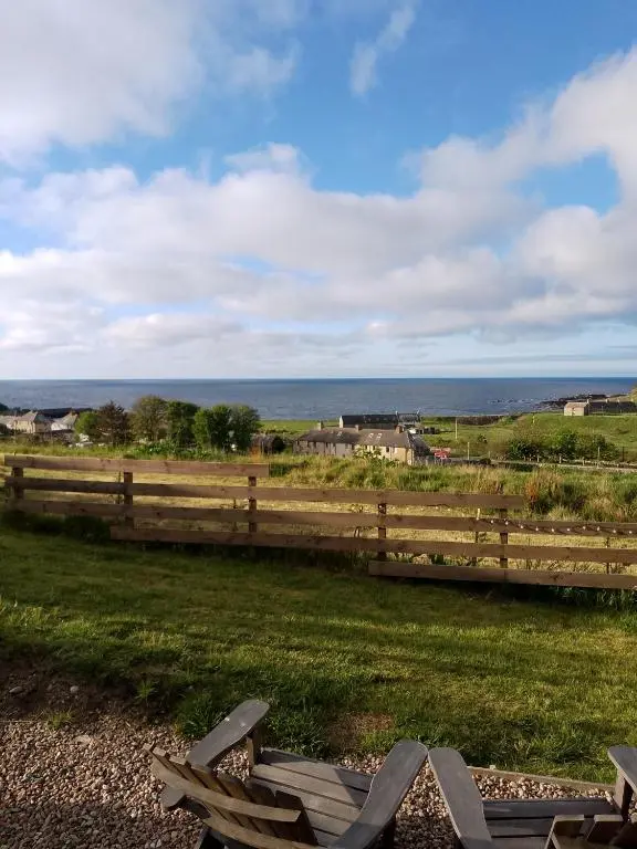a couple of wooden chairs in a field with the ocean at Camhanaich 'Half light of dawn' in Dunbeath