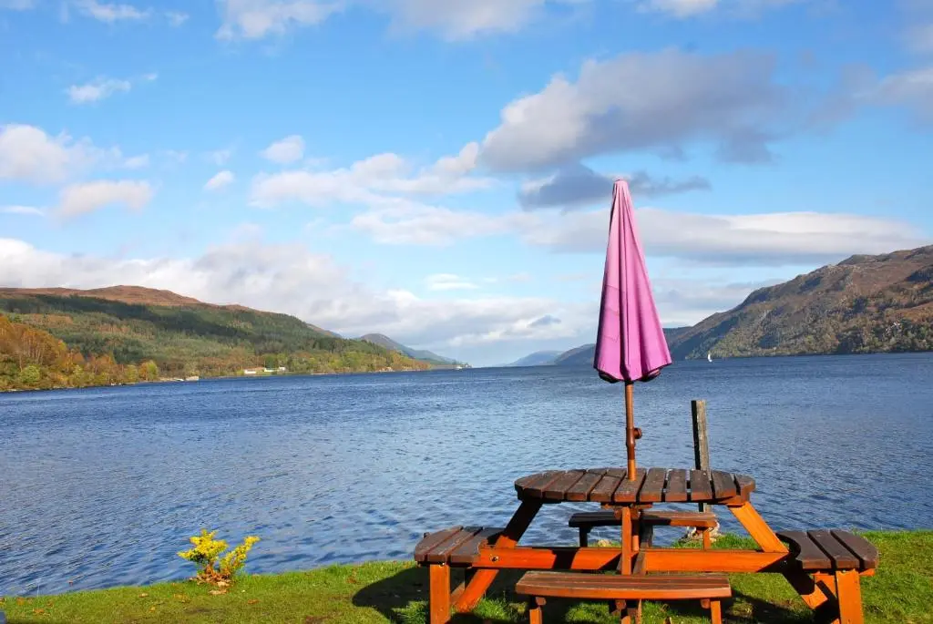 a table with an umbrella next to a lake at Highland Club Direct in Fort Augustus