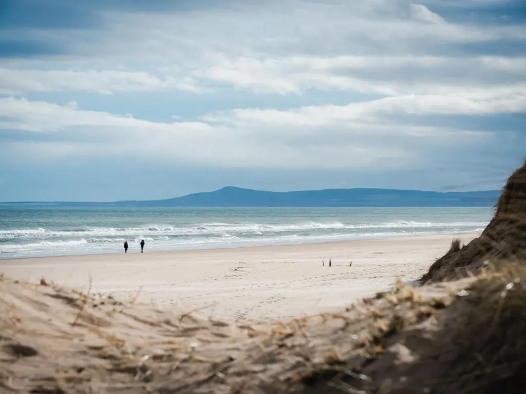 two people walking on a beach near the ocean at Apartment Harbour Nights Deluxe 7E by Interhome in Lossiemouth
