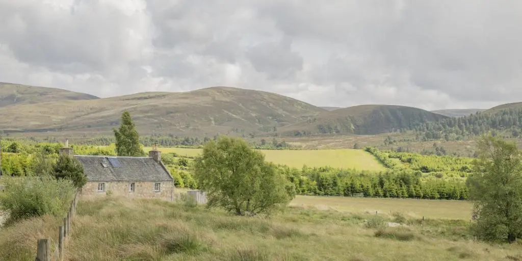 an old house in a field with hills in the background at Ellon Cottage in Nethy Bridge