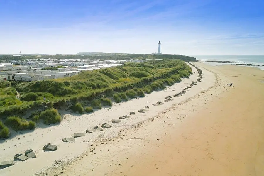 an aerial view of a beach with a lighthouse in the distance at Whispering Dunes - Silver Sands Holiday Park in Lossiemouth
