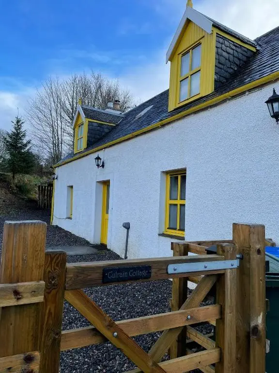 a white house with yellow windows and a wooden fence at Culrain Cottage in Fort Augustus