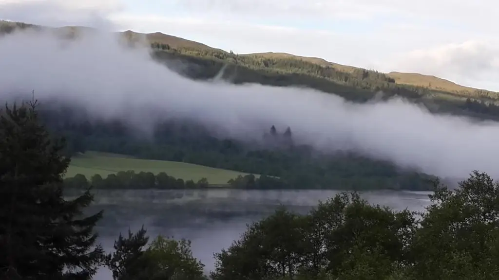 a misty lake with trees and mountains in the background at High March in Drumnadrochit