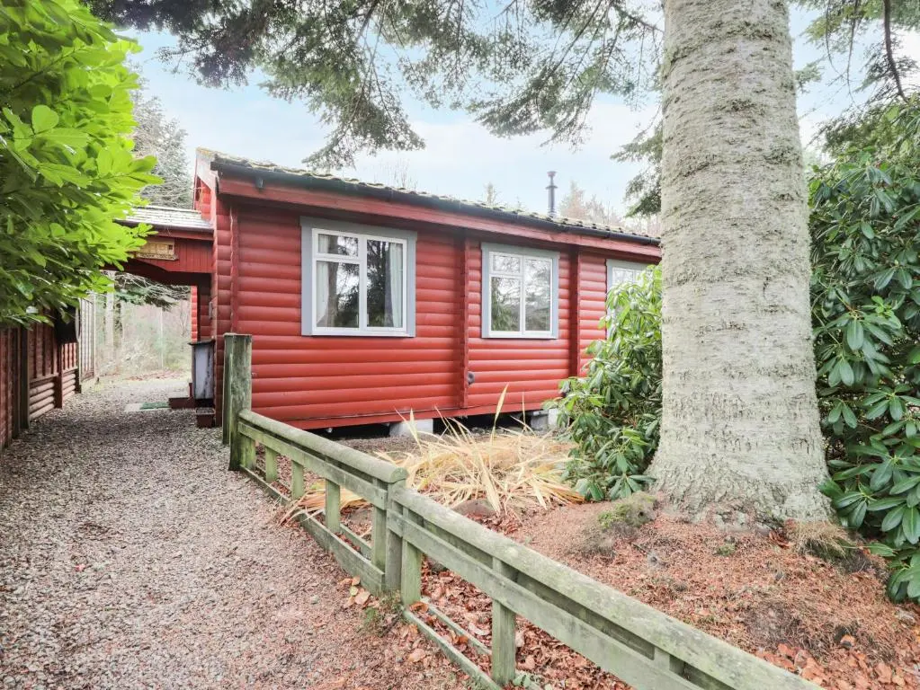 a red cabin with a fence next to a tree at Rowan Lodge in Strathpeffer