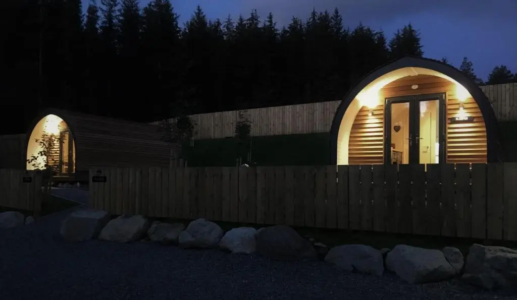a small wooden house with lights on a fence at night at Tomatin Glamping Pods in Inverness