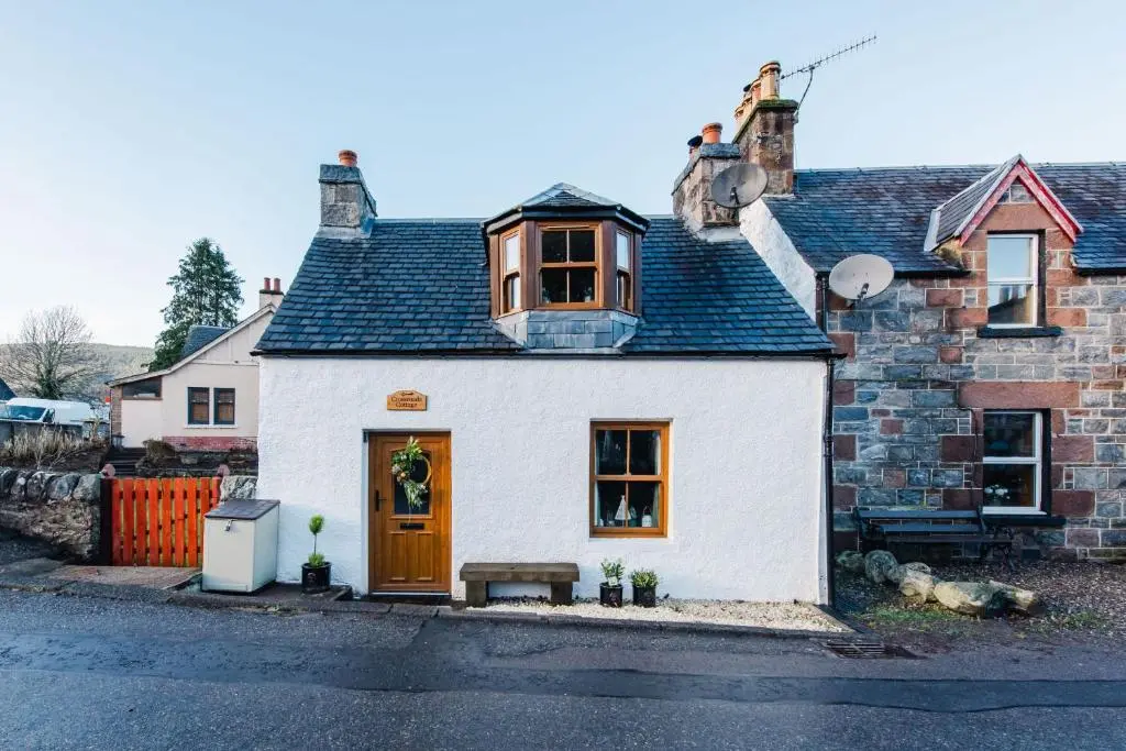 a white house with a brown door on a street at Crossroads Cottage in Drumnadrochit