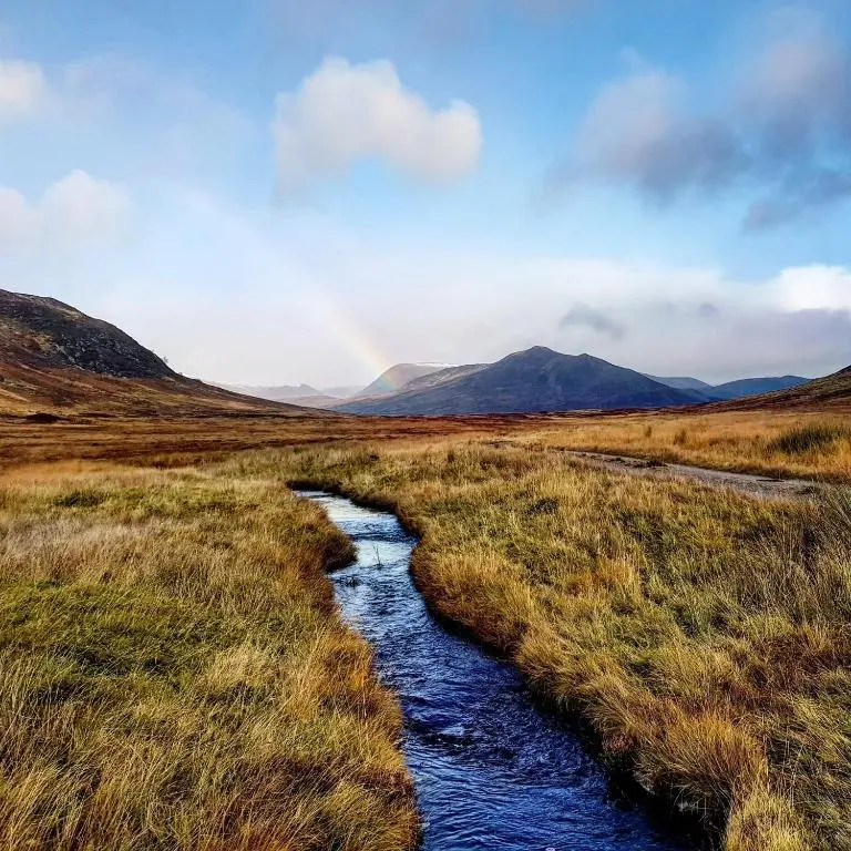 a stream in a field with a rainbow in the background at Baltinna West - by Where Stags Roar in Newtonmore